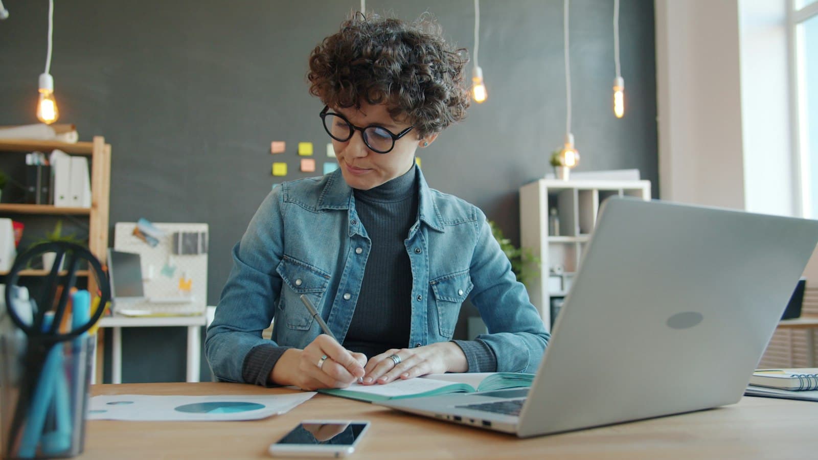 A woman writes notes in a notebook next to her laptop.